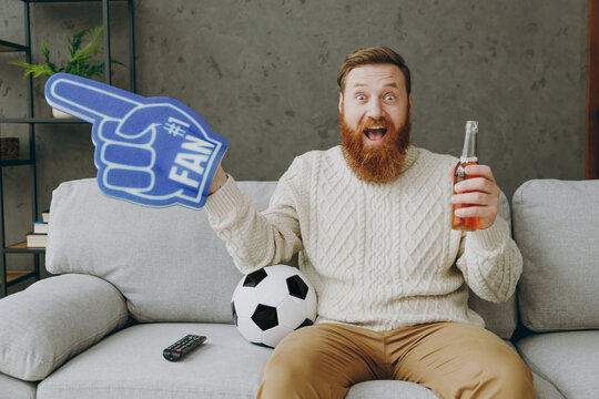 Young fun man fan wear casual clothes cheer up support football team sit on grey sofa with soccer ball bottle of beer ale foam 1 fan glove finger up rest watch tv indoor room gray wall. Sport concept.