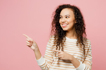 Young woman of African American ethnicity she wear light casual clothes point index finger aside indicate on workspace area copy space mock up isolated on plain pastel pink background studio portrait.