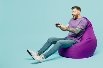 Full body fun young man he wears purple t-shirt sit in bag chair hold in hand play pc game with...