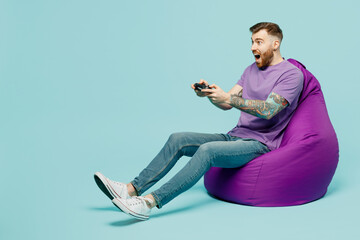 Full body fun young man he wears purple t-shirt sit in bag chair hold in hand play pc game with joystick console isolated on plain pastel light blue cyan background studio portrait. Lifestyle concept. © ViDi Studio