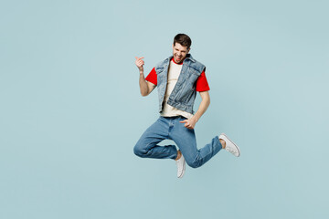 Full body singer rocker young man he wear denim vest red t-shirt casual clothes jump high do play guitar gesture isolated on plain pastel light blue cyan background studio portrait. Lifestyle concept.