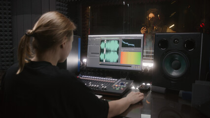 Male singer records song in soundproof room. Female audio engineer, producer uses control surface. Computer screen showing DAW software user interface with song tracks. Work in sound recording studio.