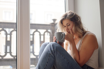 Fototapeta premium Portrait of young pretty woman sit with closed eyes on floor near window, holding grey cup, covering mouth with hand.