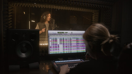 Audio engineer works with singer in sound recording studio. Vocalist in headphones sings in...