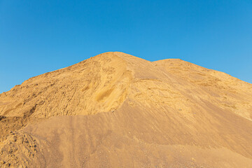 Fototapeta na wymiar A high mountain of sand in a quarry against a blue sky. Stock of bulk building materials. A big pile of sand and gravel. Photo in high resolution. A quarry for sand extraction in the photo. Sand hill.
