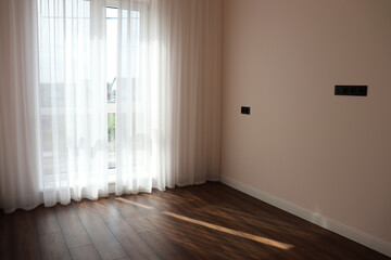 An empty room with white curtains on a large window, sunlight streaming in and creating soft shadows on the wooden floor. The space feels bright, simple, and peaceful.