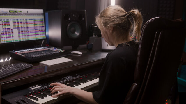 Female sound engineer, musician uses MIDI controller, digital electric piano for creating music. Computer screen shows DAW software interface with sound tracks. Recording studio. Music production.