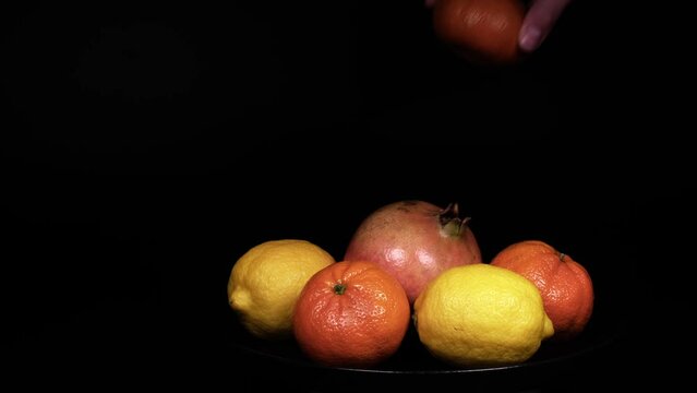 Close-up of female hand taking away fresh organic lemons, pomegranates and mandarins or tangerines piece after piece from a black plate. Beautiful citrus fruits against isolated black background.