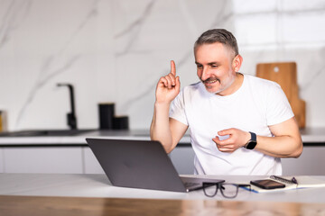Smiling middle aged caucasian male with beard in white t-shirt eats sandwich, has video call,...