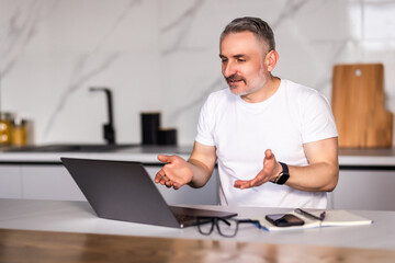 Smiling middle aged caucasian male with beard in white t-shirt eats sandwich, has video call, gesticulates in kitchen interior.