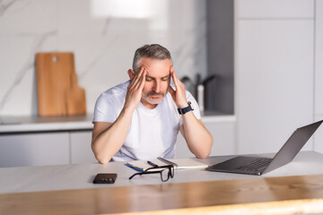 Busy mature man with beard in glasses works with documents behind laptop in minimalist kitchen interior. Home bookkeeping, finance, paying mortgage, loans and taxes bills, free space