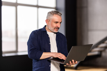 Mature man holding using laptop standing leaning on white wall, posing looking at camera.