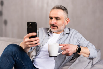 Man drinking coffee and using mobile phone at home. Adult caucasian male enjoying morning routine at living room sofa