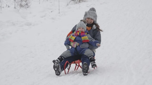 Young mother and happy child on sleigh sliding on snow slope in winter