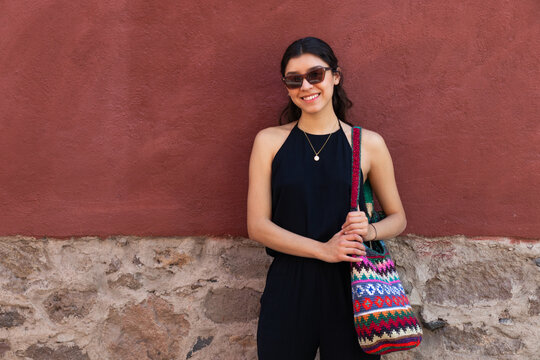 Young woman with colorful handbag standing by red wall