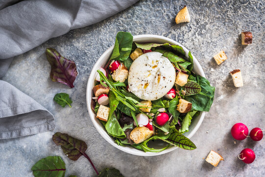 Mixed salad with radishes, homemade croutons and burrata cheese on gray background, top view