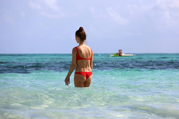 Slim girl wearing red bikini going to swim in blue sea water. Beach vacation on tropical coast