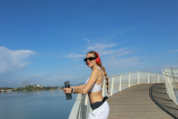 Fototapeta na wymiar Fit woman in her 30s standing on the bridge with the water bottle, wearing white sportswear and red headphones. Background, copy space for text, close up.
