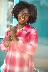 Half body portrait of latin girl with afro hairstyle and glasses holding a takeaway coffee outdoors with happy expression dressed in casual clothes, isolated on a vibrant green background.