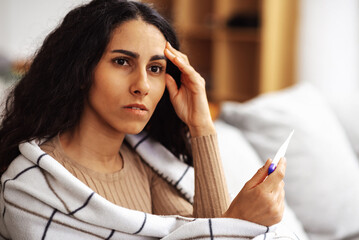 Young beautiful Arab woman falls ill and looking ahead thoughtfully. Woman holds a thermometer with one hand and she holds her head with her other hand with the other while sitting at home.