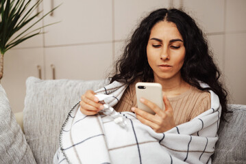 Beautiful young arab woman sitting on the couch in her room wrapped in a blanket. Holds a smartphone in one hand and an electronic thermometer in the other pill. She dials the family doctor's phone.