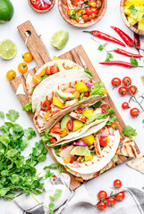 Tacos with grilled chicken fillet, salsa sauce, mango, cilantro and red onion in corn tortilla on cutting board. White table background, top view