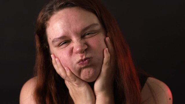 Portrait of middle-aged woman with long red hair puffing blowing cheeks, holding hands on cheeks, pinching, grimacing, shaking head, having fun. Facial exercises, self-treatment, massage, skincare.