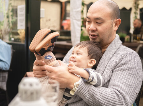 Flexible male infant smiling sitting on his dad's legs holding his son's legs, kid playing with father in beer garden at pub. Joyful lifestyle. Happy memories. Multicultural family, mixed race.