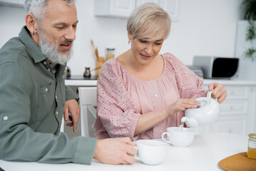 joyful woman pouring tea near bearded husband smiling in kitchen.