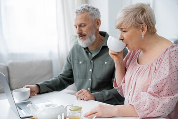 positive middle aged couple looking at laptop while enjoying morning tea in kitchen.