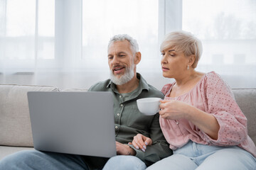 middle aged woman holding tea cup while watching film on laptop near smiling husband on sofa in living room.