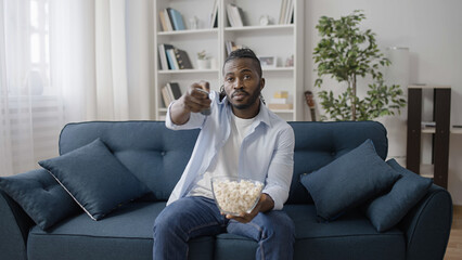 African American man sitting on sofa, watching movie on TV and eating popcorn
