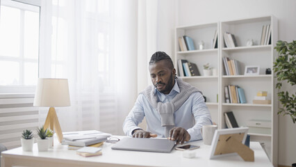 An African American man is sitting at his remote workspace with his laptop at home