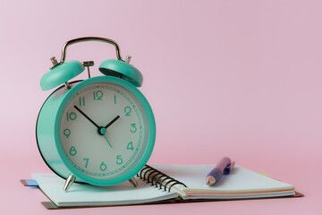 A bell alarm clock with a notebook and a pen on pink background for the concept of time management for work and study.