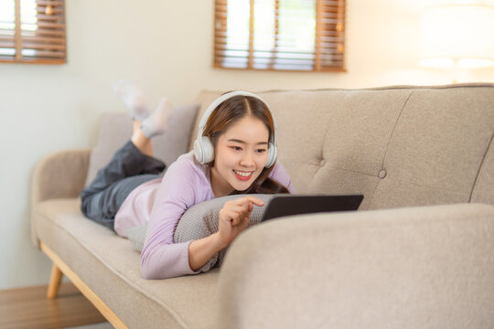 Happy young teenage beautiful Asian woman using tablet for home entertainment  while sitting on the couch in the house.