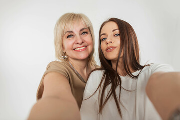 Beautiful elderly cheerful mother with a smiling happy pretty daughter takes a selfie on a smartphone on a white background in the studio