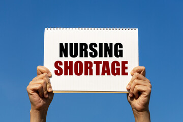 Nursing shortage text on notebook paper held by 2 hands with isolated blue sky background. This...