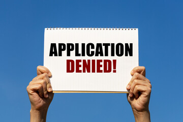 Application denied text on notebook paper held by 2 hands with isolated blue sky background. This...