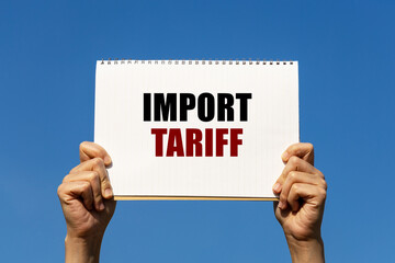 Import tariff text on notebook paper held by 2 hands with isolated blue sky background. This...