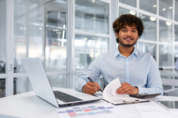 Portrait of young boss inside office at workplace, successful hispanic businessman smiling and looking at camera, man signing contracts and invoices, financier smiling and looking at camera.