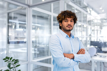 Fototapeta premium Portrait of a successful young hispanic programmer standing in a modern office with his arms crossed on his chest and looking confidently at the camera.
