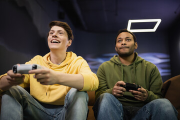 KYIV, UKRAINE - FEBRUARY 13, 2023: Low angle view of cheerful man playing video game with indian friend in gaming club.