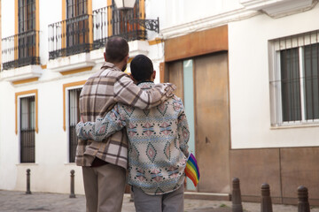 Real marriage of gay couple, hugging on the street, with a gay pride flag in their pocket and seen from the back. Concept lgtb, lgtbiq+, couples, in love, pride, hugging.