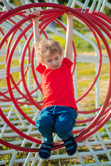 Fototapeta premium A curly-haired little boy in a red T-shirt walks on the playground. The child develops in the process of sports play. Jumping on ropes and horizontal bars