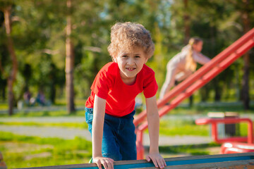 Fototapeta na wymiar A curly-haired little boy in a red T-shirt walks on the playground. The child develops in the process of sports play. Jumping on ropes and horizontal bars