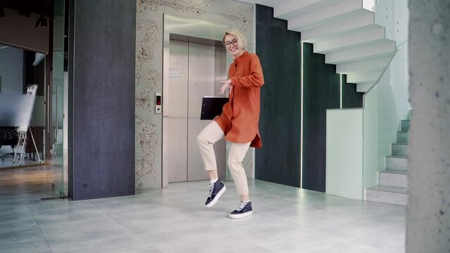 Happy joyful young and attractive woman dancing victory dance in corridor or hall of modern business center. Female office worker cheerfully celebrating achievement. Victory dance of a funny employee
