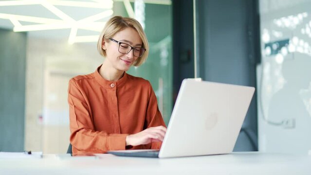 Happy young employee wearing glasses typing on laptop while sitting at workplace in modern office. A smiling blonde female chats online, communicates with a colleague, a friend, replies to a client