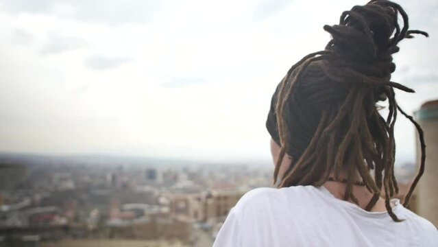 a lonely young man with dreadlocks gathered in a bun looks at the city from a high place. Looking behind. a young Brazilian man with dreadlocks and an earring in his ear looks at the city landscape