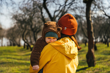 a mother woman in a yellow raincoat in a red hat stands with her back and holds a child in a teddy bear in her arms