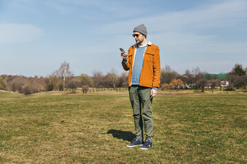 a man in a hat and an ocher corduroy jacket stands in a field in a park with a phone in his hands...